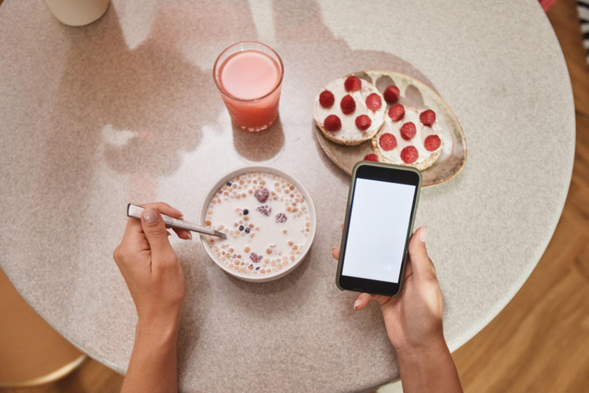 Top-down view of a breakfast: bowl of cereal with berries, raspberry toast, a pink drink, and a person using a smartphone.